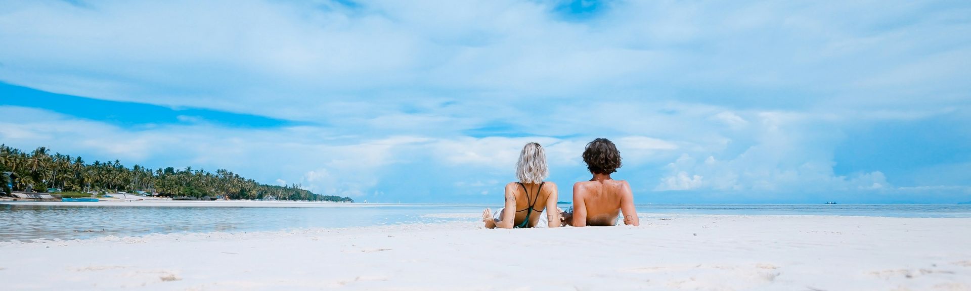 Besondere Urlaubsziele: Ein Paar liegt in Badesachen auf dem Rücken am weißen Sandstrand und blickt aufs Wasser