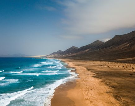 Fuerteventura im Dezember: Ein breiter und langer Sandstrand an einer wüstigen Vulkanlandschaft am blauen, wilden Meer