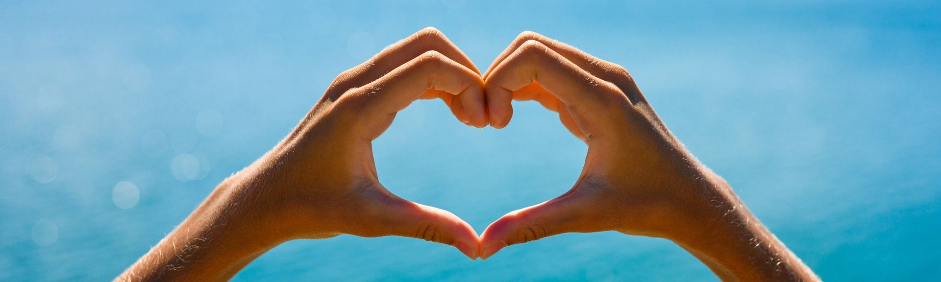 Zwei Hände formen ein Herz und dahinter glitzert das blaue Meer hindurch