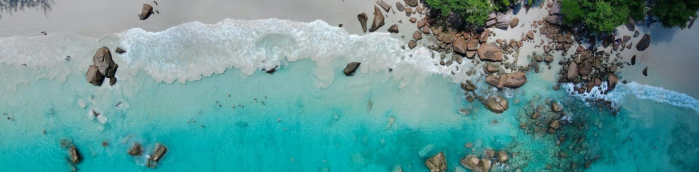 Alle Urlaubsziele: Eine Luftaufnahme von einem türkis, glasklaren Wasser, leichte Wellen schlagen an einem Sandstrand mit Steinen.