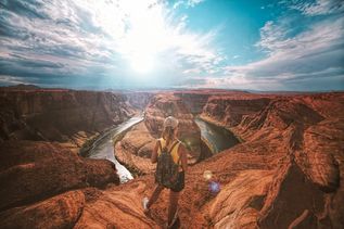 Eine Frau mit Rucksack und Cap steht vor einem riesigen Canyon unter hellblauem Himmel mit Schleierwolken.