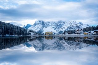 Winterurlaub Italien: Ein spiegelglatter Bergsee mit Häusern in einer verschneiten Landschaft