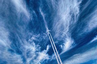 Am tiefblauem Himmel mit dünnen Wolkenschleiern, fliegt ein weißes Flugzeug, welches hinter sich zwei weiße Kondenzstreifen her zieht.