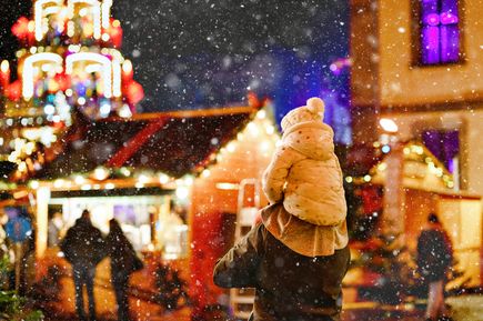 Eine Person trägt ein kleines Kind auf den Schultern über einen verschneiten, leuchtenden Weihnachtsmarkt