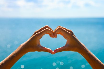 Frühbucher 2026: Zwei Hände formen ein Herz und im Hintergrund glitzert das blaue Meer durch