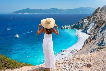 Eine Frau mit Sonnenhut und weißem Kleid steht an einem Abhang und blickt auf einen weiß-grauen Strand und das Meer