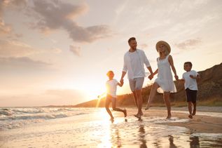 Osterurlaub: Eine Familie mit zwei Kindern laufen Hand-in-Hand am Strand entlang, die untergehende Sonne im Rücken.