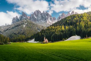 Italien Urlaub: Spitze, hohe Berge ragen hinter einer Wiese mit einer idyllisch gelegenen Kirche empor.