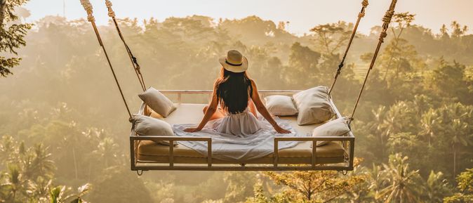 Eine Frau mit Sonnenhut sitzt in einem hängenden Bett mitten im Djungel. Fernreisen im Winter.