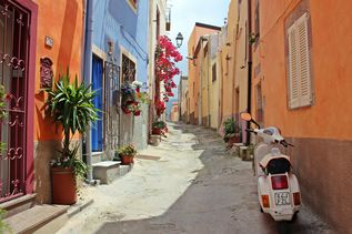 Eine mediterrane Gasse mit Bunten Häusern, einer Vespa und Blick aufs Meer.