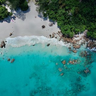 Eine Luftaufnahme von einem türkisen, glasklarem Meer, welches am weißen Strand angrenzt. Einige graue Steine befinden sich im Wasser und am Strand.