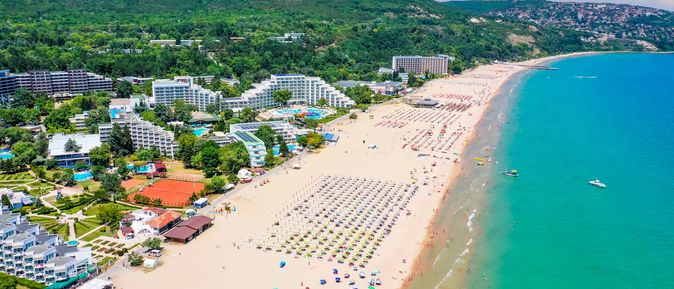 An einem langen, goldenen Sandstrand mit türkisem Meer stehen viele Sonnenliegen mit Schirmen. Angrenzend eine grüne Landschaft mit großen Hotelanlagen.