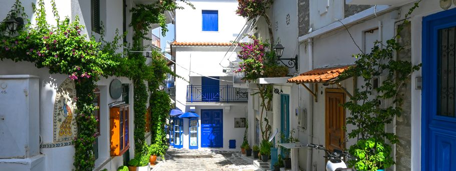 Eine schmale Gasse mit Kopfsteinpflaster führt zwischen weißen Häusern mit blauen Türen und Fensterläden.