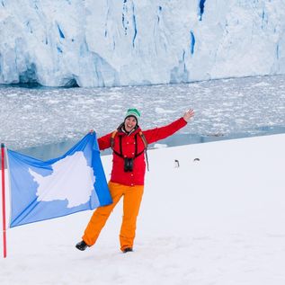 Oceanwide Expeditions: Eine Frau in Winterkleidung steht auf einer schneebedeckten Fläche und hält eine Flagge mit einem Landumriss fest.e