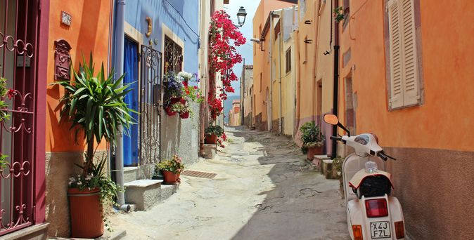 Italien Urlaub: Eine schmale Gasse zwischen bunten Häusern mit mediterranen Blumen und einer italienischen Vespa.