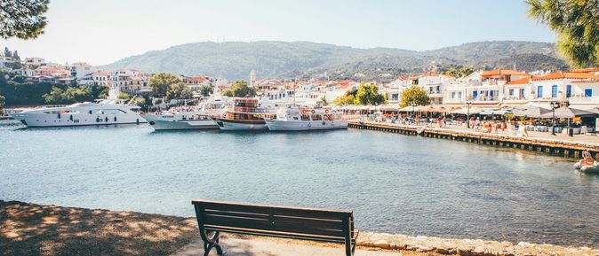 An einem hübschen, kleinen Hafen steht eine leere Bank. Im Hintergrund legen einige Boote an und eine mediterrane Stadt erstreckt sich.
