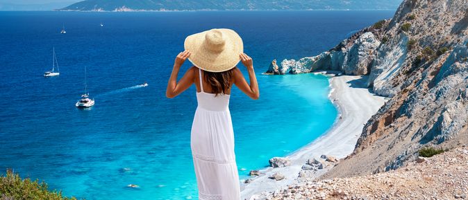 Eine braungebrannte Frau mit weißem Sommerkleid und Sonnenhut steht an einem Felsabhang. Sie blickt auf einen grau-weißen Strand, ein türkis-tiefblaues Meer mit 4 Segelbooten und im Hintergrund eine Bergkette.