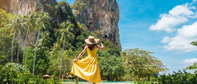 All inclusive Thailand: Eine Frau im gelben Kleid und Sonnenhut steht vor Palmen und riesigen Kalksteinfelsen