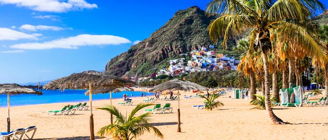 Osterurlaub: Ein Strand mit Sonnenliegen und Palmen vor einem Berg mit bunten Häusern