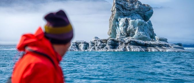 Oceanwide Expeditions: Eine Person in roter Jacke und Mütze befindet sich auf einem Schiff und blickt auf einen skurrilen Felsen im Wasser