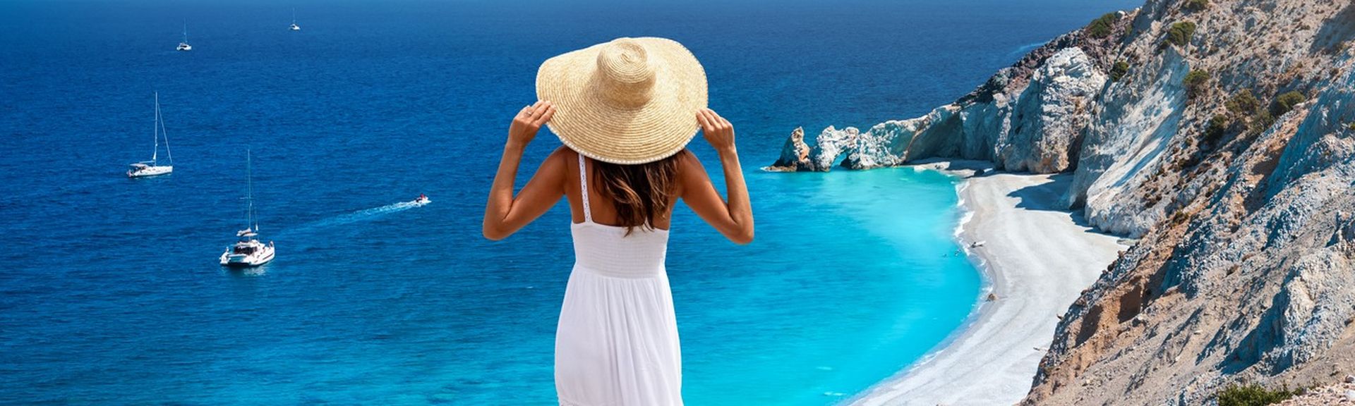 Eine Frau im weißen Sommerkleid mit Hut steht am Abhang und blickt auf eine Bucht mit weiß-grauem-Strand und türkis-blauem Wasser.