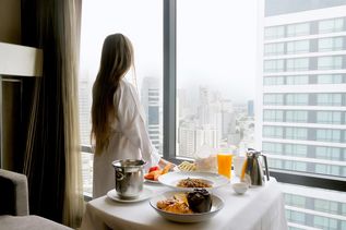 Eine Frau in weißem Bademantel und braunen Haaren steht am Fenster eines Hotels und blickt hinaus. Neben ihr steht ein gedeckter Frühstückstisch ebenfalls mit weißer Tischdecke.