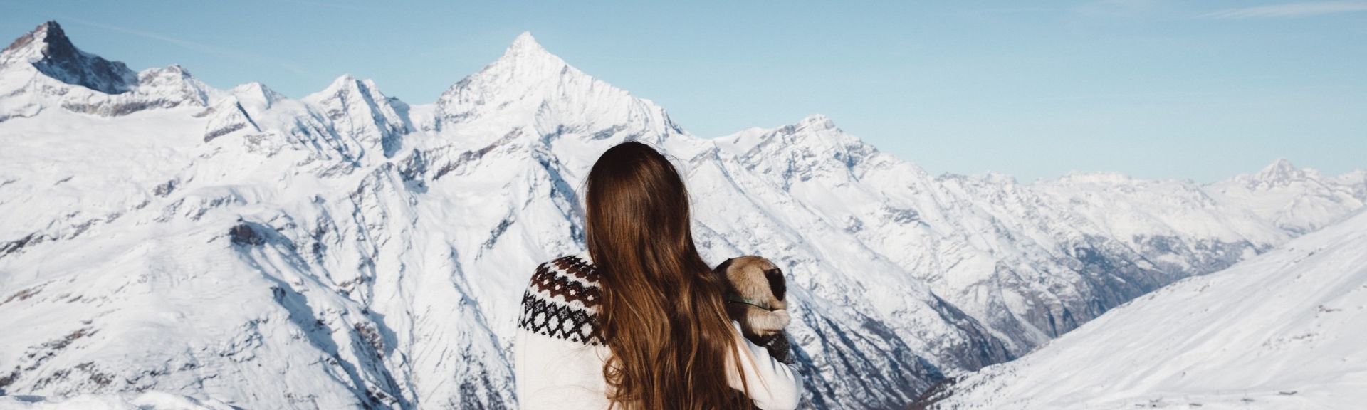 Winterurlaub in den Bergen: Eine braunhaarige Frau sitzt mit Norwegerpulli und ihrem Hund auf einem verschneiten Berggipfel
