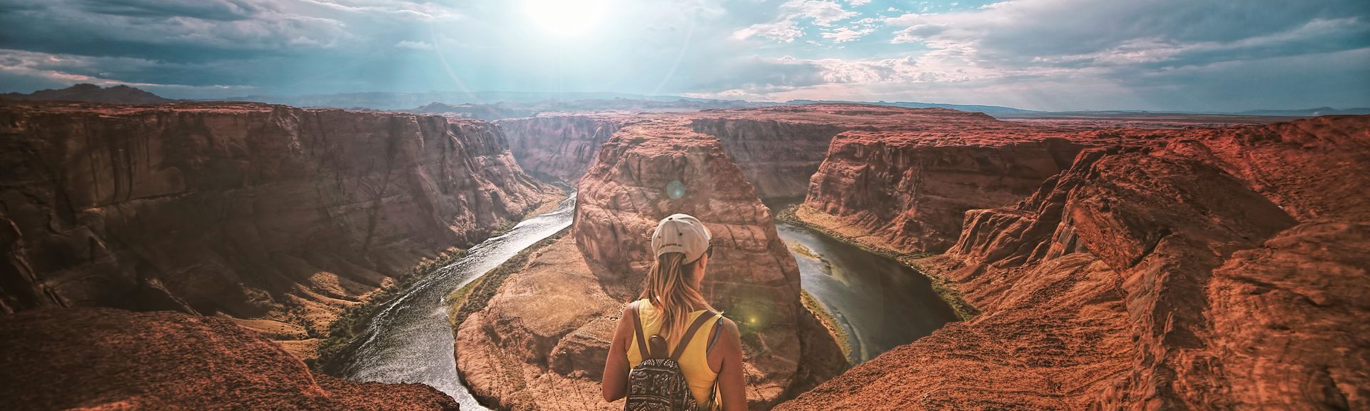 Eine Frau mit Rucksack und Cap steht vor einem riesigen Canyon unter hellblauem Himmel mit Schleierwolken.