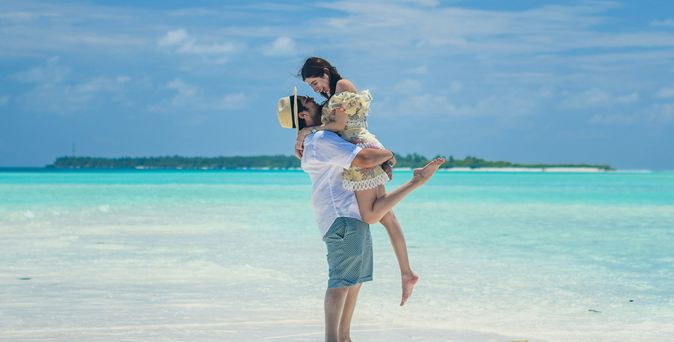 Adults only Urlaub: Ein Mann hält eine Frau im Arm, die beiden stehen am seichten Wasser eines türkis-blauen Meers.