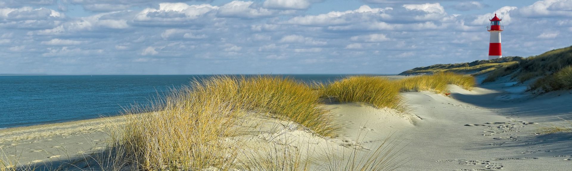 Urlaub in DE: Ein weißer Strand mit Dünengras und einem rot-weißen Leuchtturm.