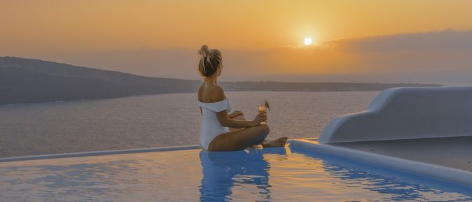 Eine Frau in weißer Badekleidung sitzt im Schneidersitz an einem Infinity-Pool und blickt in den Sonnenuntergang.