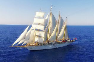 Ein schönes, riesiges Segelschiff mit mehreren Segelmasten und weißen Segeln gleitet auf dem dunkelblauen Meer unter hellblauem Himmel dahin.