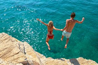 Ein Junge und ein Mädchen halten sich an den Händen und springe von einer Felsklippe ins Meer.