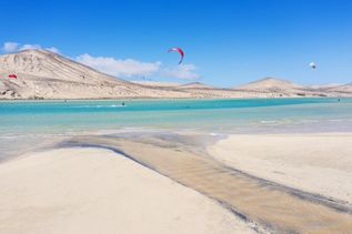 Frühbucher: Ein weißer, feinsandiger Strand mit türkisem Wasser und einigen Kitesurfern an einer kargen Wüstenlandschaft