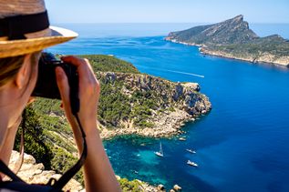 Eine Person mit Sonnenhut blickt durch ihre Kamera auf eine Bucht zwischen zwei begrünten Berglandschaften auf Mallorca.