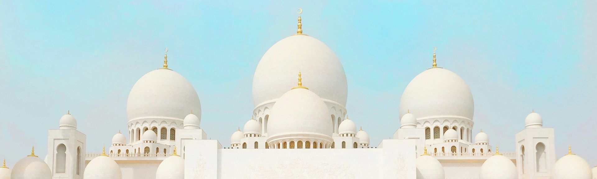 Urlaub im Orient: Eine prächtige, weiße Moschee mit mehreren Runden Kuppeln unter hellblauem Himmel und goldenen Ornamenten