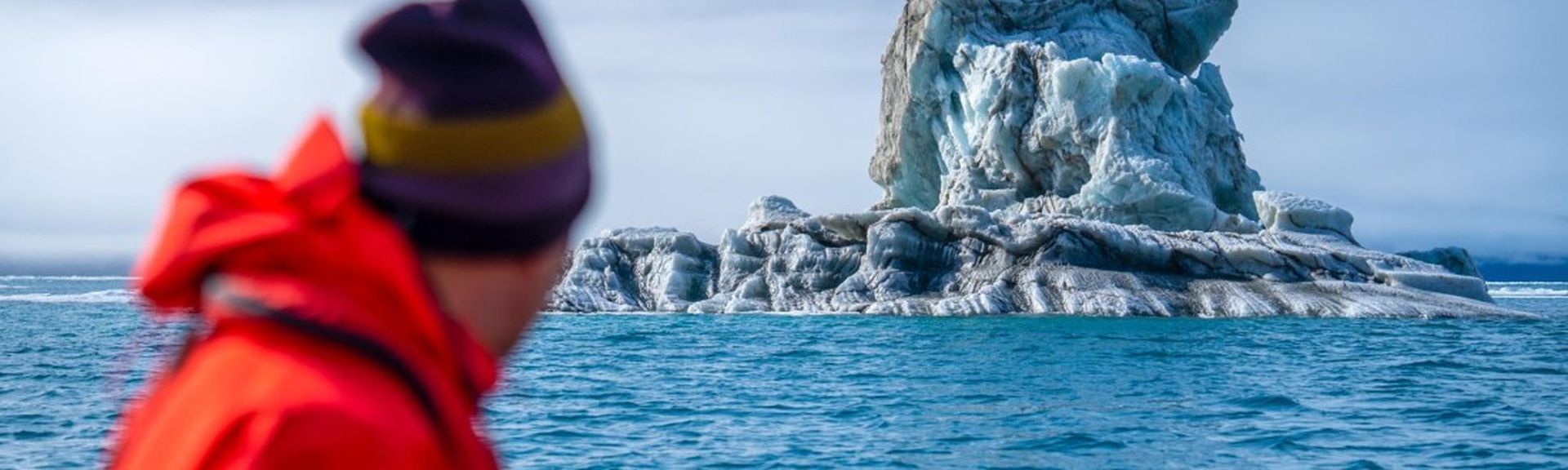 Oceanwide Expeditions: Eine Person in roter Jacke und Mütze befindet sich auf einem Schiff und blickt auf einen skurrilen Felsen im Wasser