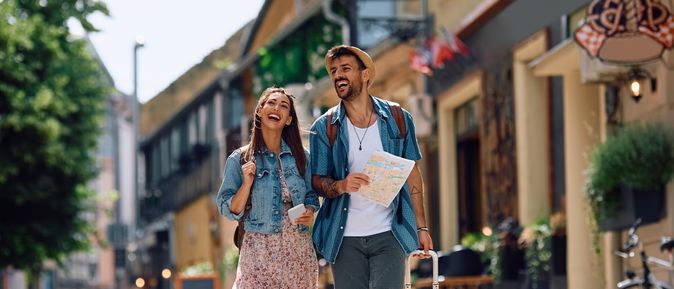 Städtereisen: Ein junges, lachendes Paar läuft mit Stadtplan durch eine charmante Straße