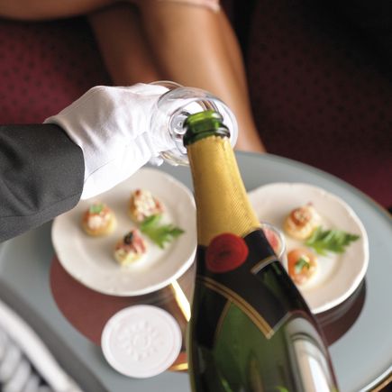 MSC Yacht Club: Eine Person mit schwarzem Blaser und weißen Handschuhen gießt Champagner in ein Glas, darunter Kanapés.