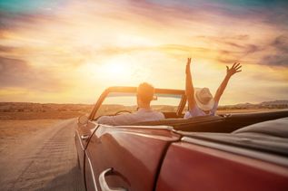 In einem roten Cabrio fahren ein Mann und eine Frau der Sonne auf einer Landstraße entgegen. Die Frau reißt die Hände fröhlich in die Luft.