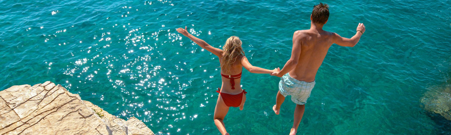 Ein Junge und ein Mädchen halten sich an den Händen und springe von einer Felsklippe ins Meer.