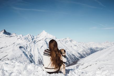 Winterurlaub in den Bergen: Eine braunhaarige Frau sitzt mit Norwegerpulli und ihrem Hund auf einem verschneiten Berggipfel
