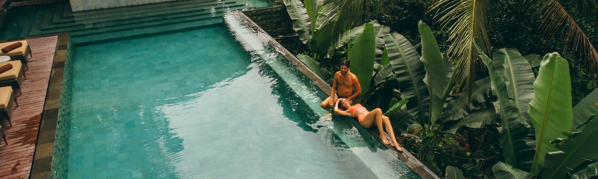 Luxus: Ein Pärchen in Badesachen liegt am Rand eines Infinitypools direkt an tropischen Pflanzen