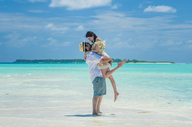 Adults only Urlaub: Ein Mann hält eine Frau im Arm, die beiden stehen am seichten Wasser eines türkis-blauen Meers.