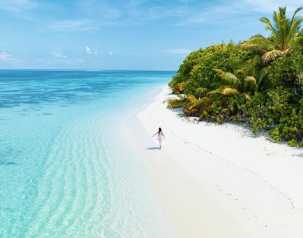 Malediven im Dezember: Eine Frau läuft an einem einsamen, weißen Strand am türkis-blauen Meer entlang.